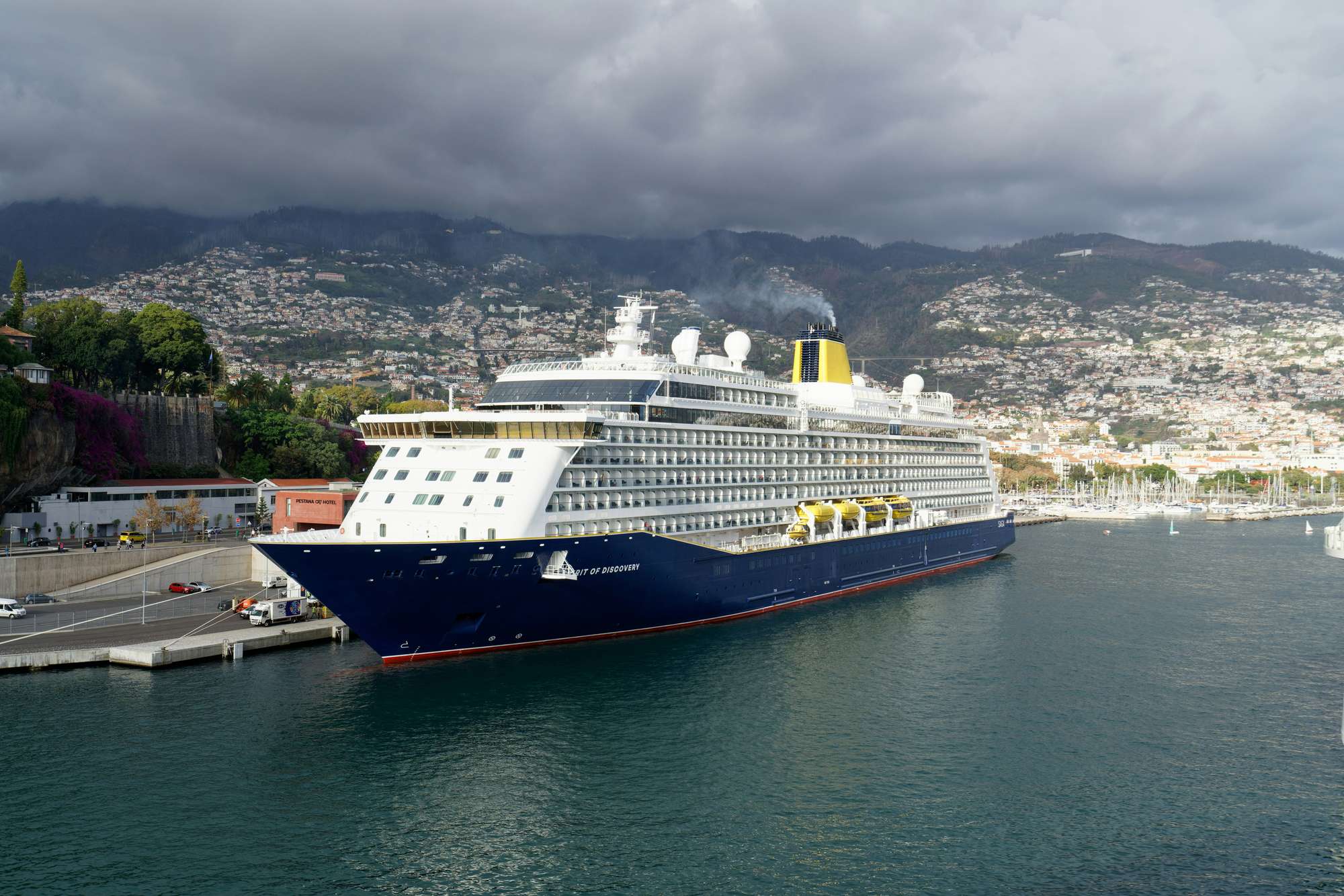 A cruise ship at the pier in Funchal, with the city climbing the hills behind it under heavy cloud