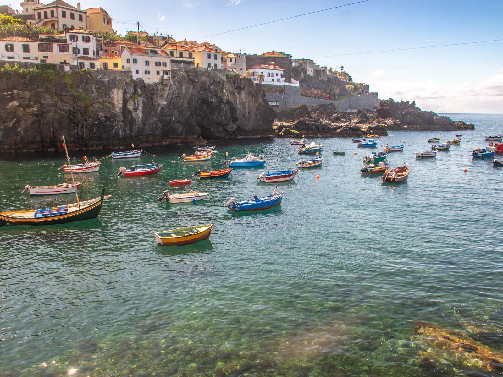 Painted fishing boats on clear green water in the harbour at Câmara de Lobos, houses on the cliff above