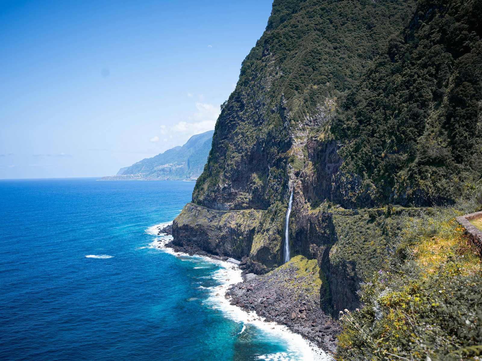 The Véu da Noiva waterfall falling down a green sea cliff onto the boulders of the north coast
